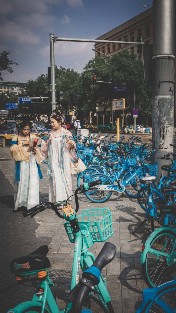 Women in traditional-style clothing walking past rows of shared bicycles on a street in Beijing, China.