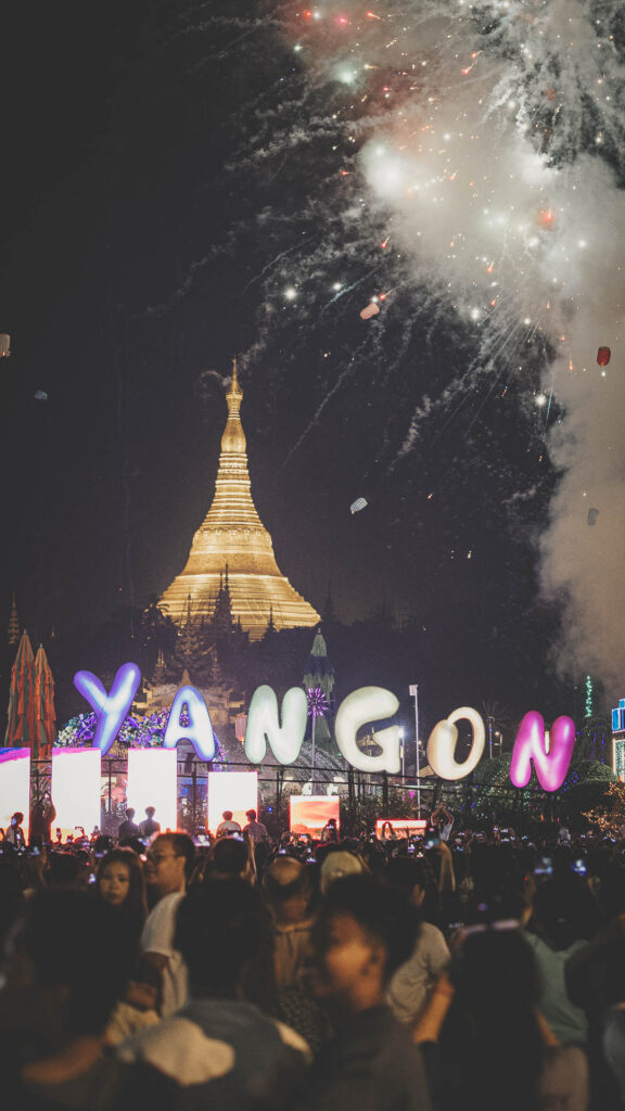 Crowds gather during a nighttime festival with fireworks and an illuminated pagoda in Yangon, Myanmar.