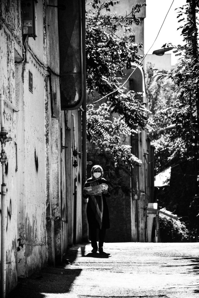 A person wearing a face mask walks along a narrow, tree-lined street in Iran.